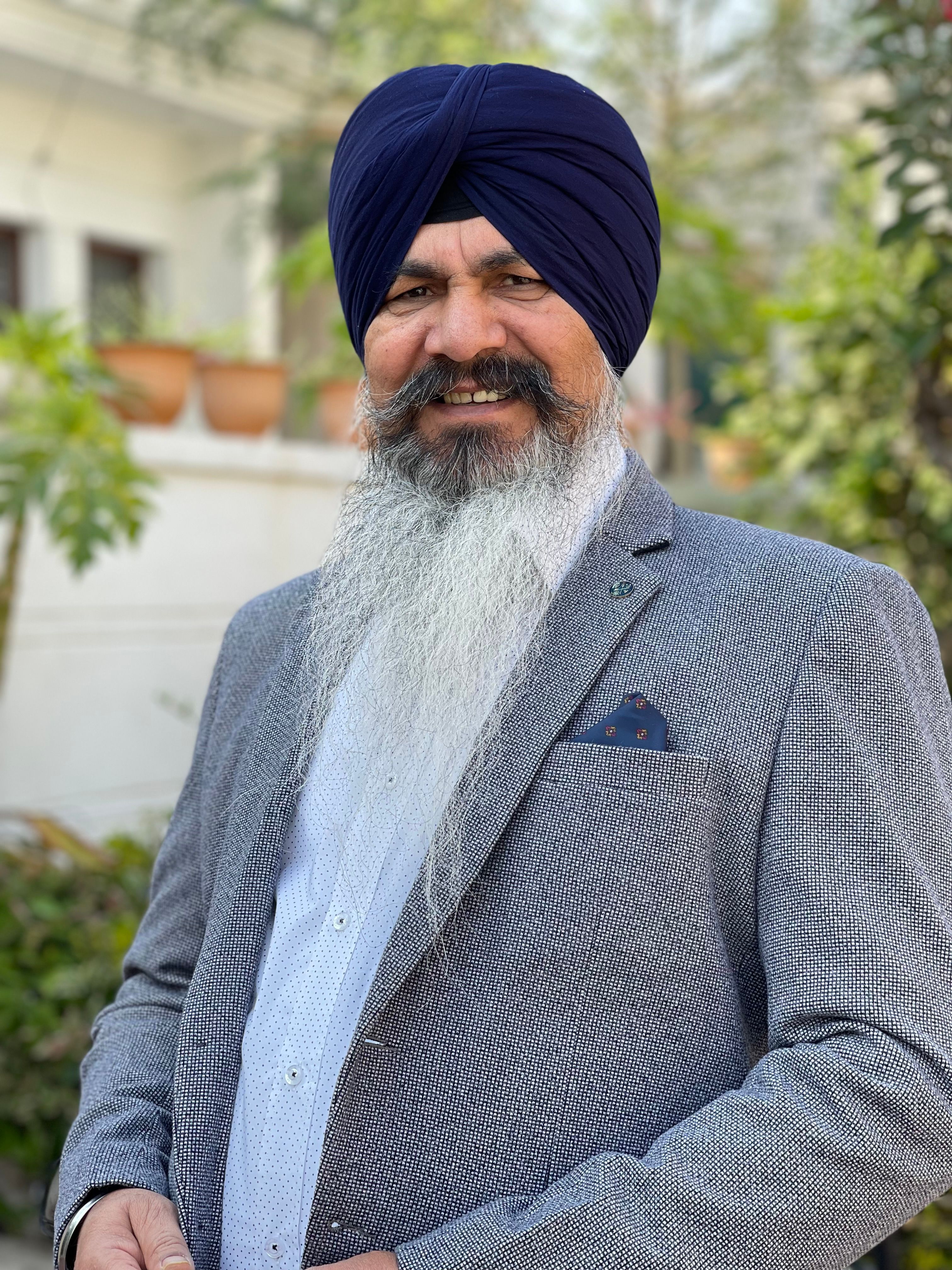 Elderly man wearing a turban and blazer smiles outdoors in a sunny setting.