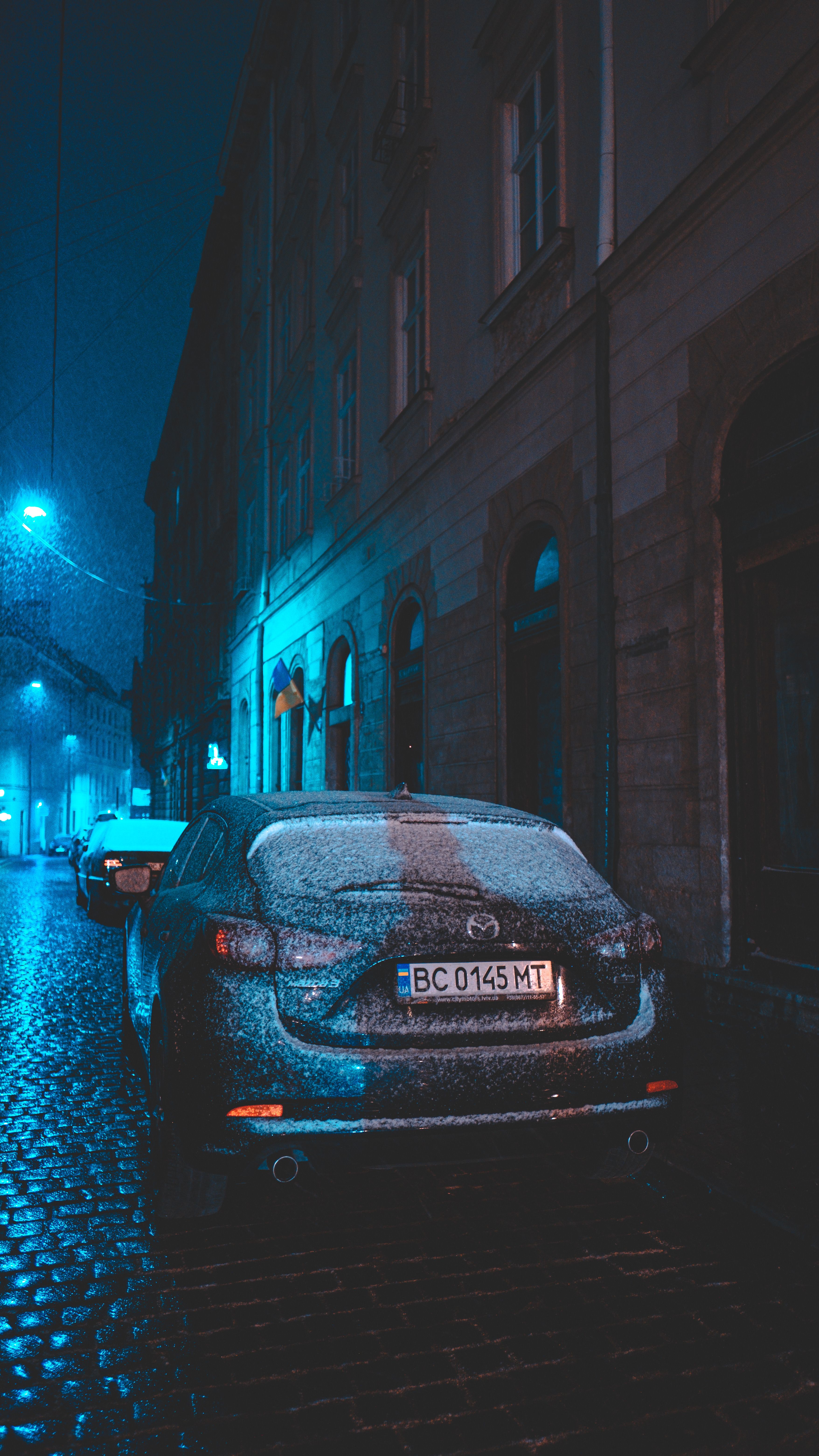 A dimly lit street with snow-covered cars parked along a cobblestone road during a winter night.