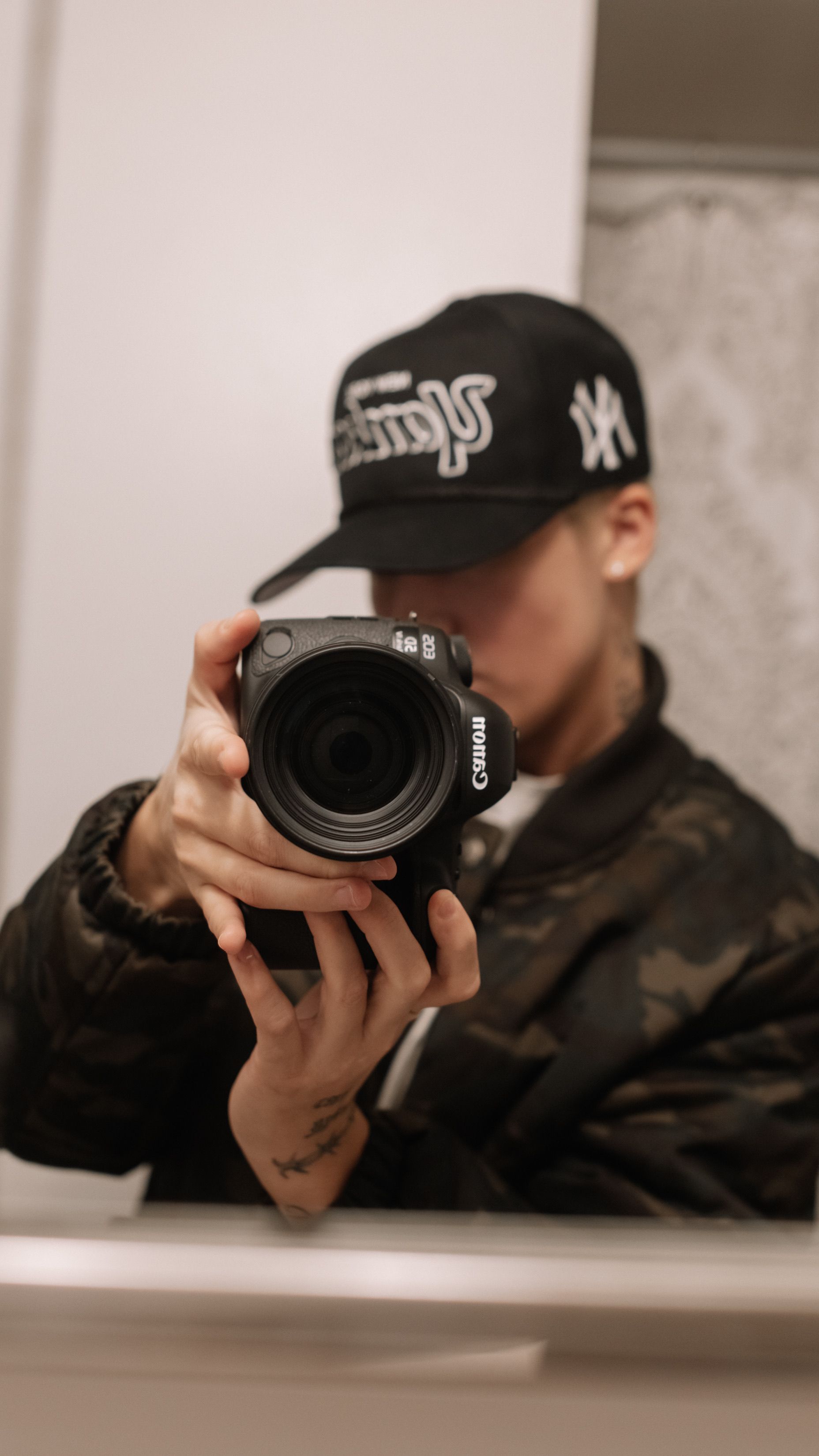 Portrait of a person taking a mirror selfie with a professional camera, wearing a stylish cap.