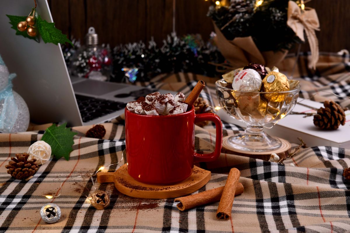 Cozy winter scene with hot chocolate in a red mug, topped with marshmallows and surrounded by holiday decorations.