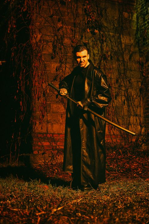 Free A man in a black coat holding a sword stands against a brick wall in a dramatic nighttime setting. Stock Photo