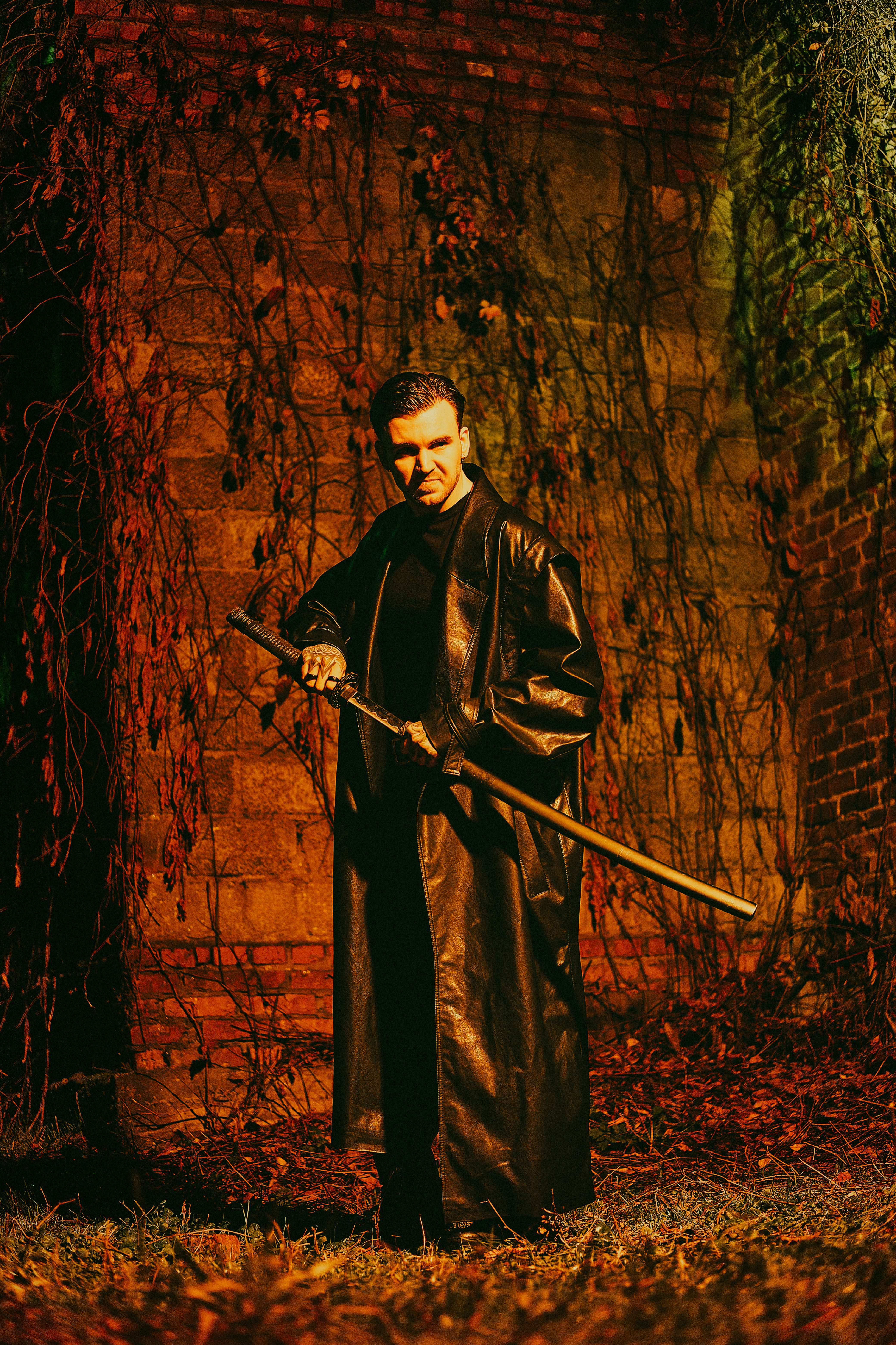 A striking image of a man in a leather coat holding a sword, standing against a rustic wall.