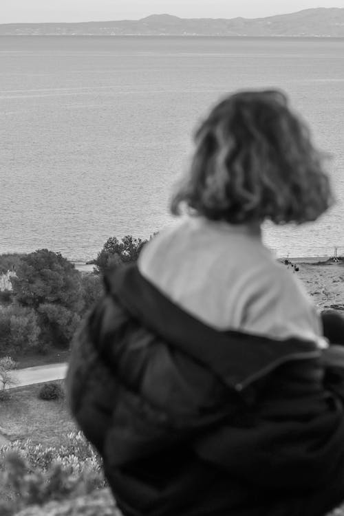 Grayscale Photo of Woman in White Shirt and Black Jacket Looking at the Ocean