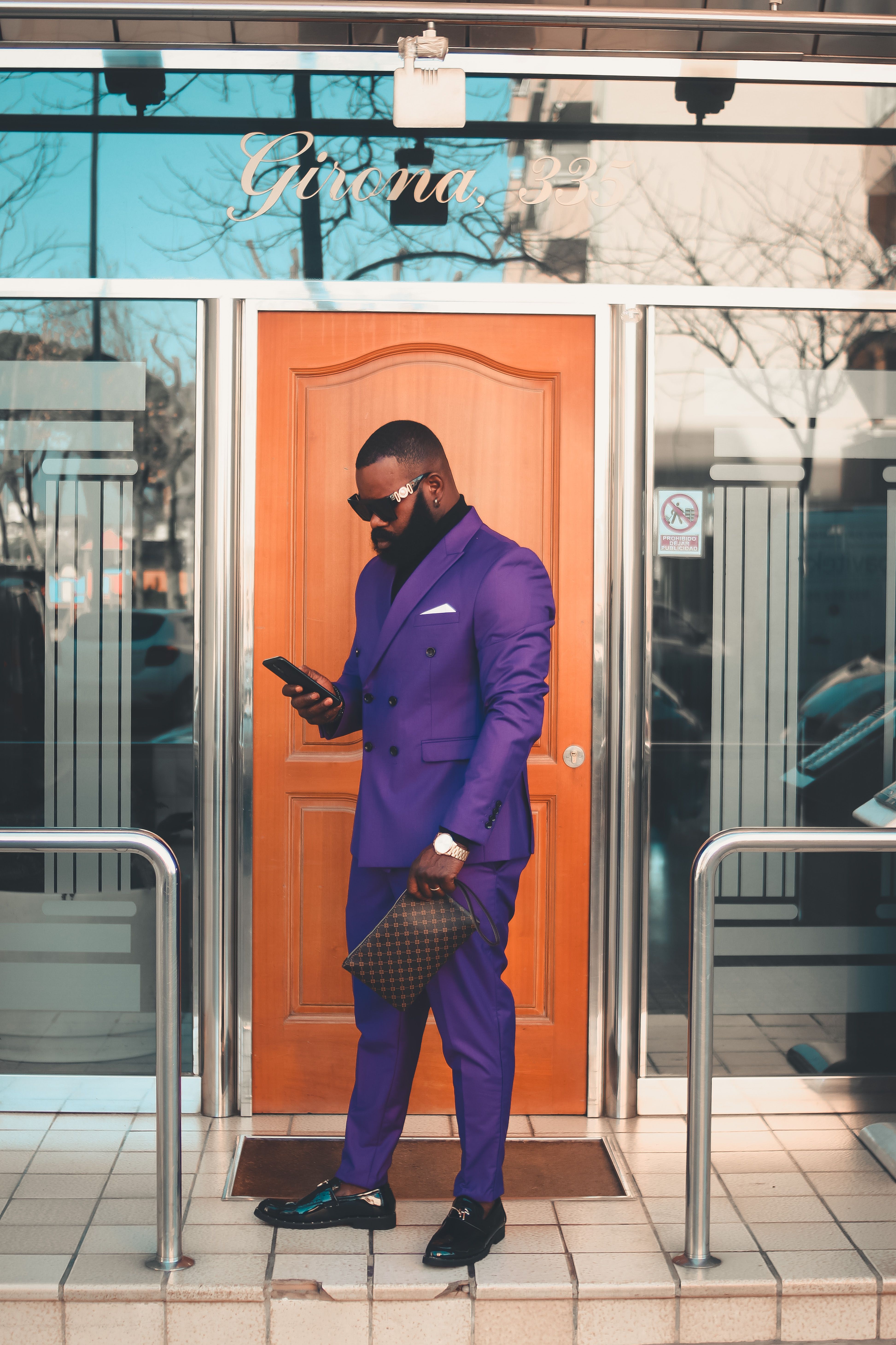 Fashionable man in violet suit stands outside building, holding a phone and clutch.