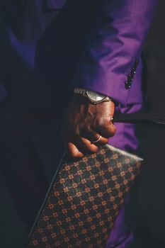 Close-up of a man's hand in a purple suit holding a fashionable clutch, exuding style and luxury.