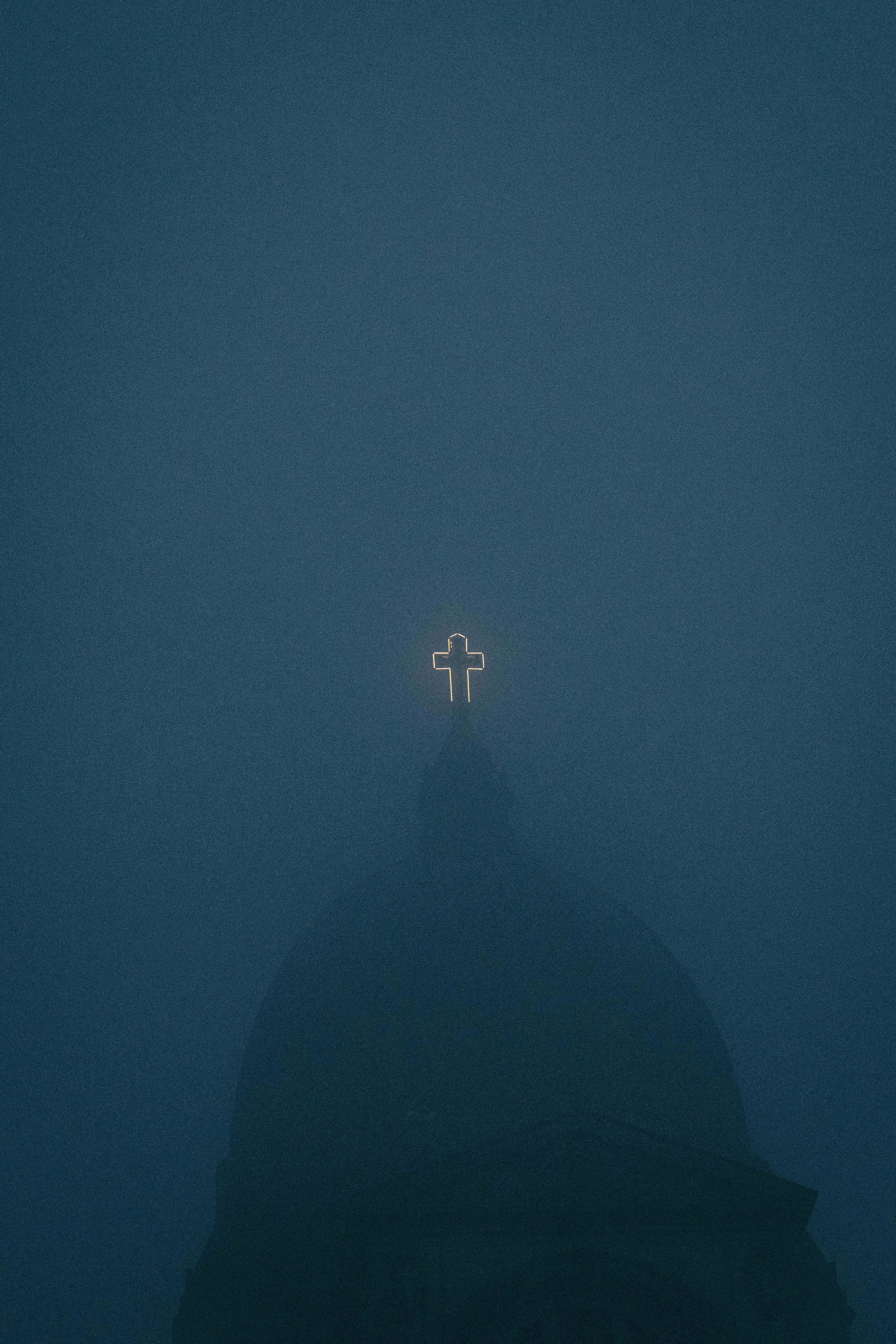 A glowing cross atop a church emerges through thick fog, creating a mystical evening atmosphere.