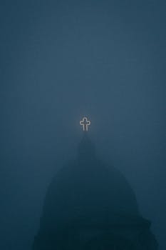 A glowing cross atop a church emerges through thick fog, creating a mystical evening atmosphere.