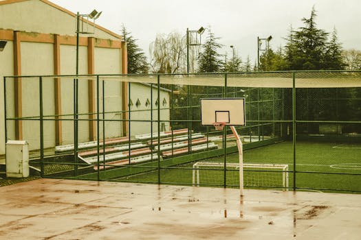 Empty outdoor basketball court with wet surface, surrounded by trees and buildings, captured during daytime.