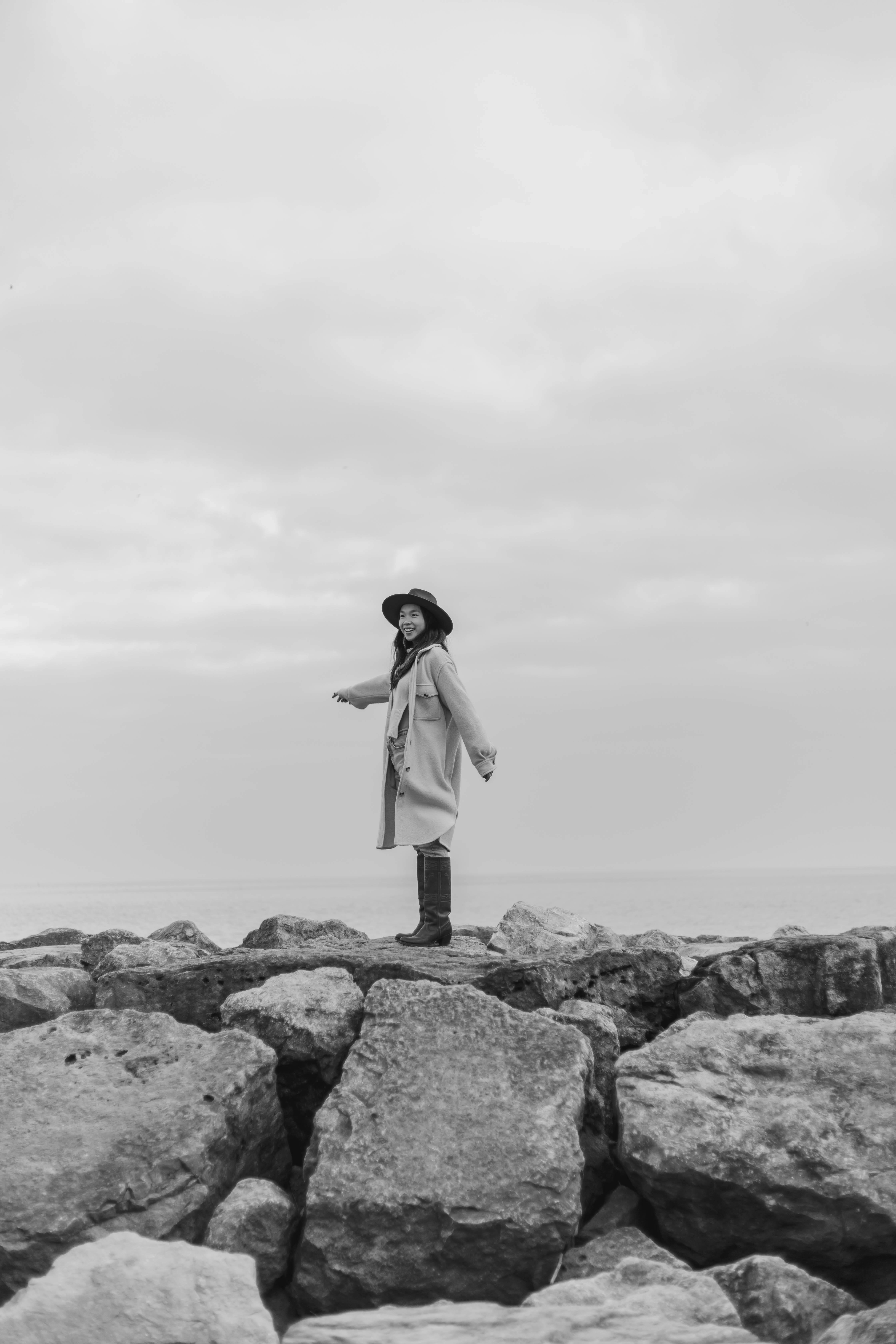 Black and white photo of a woman standing playfully on rocky shore under overcast sky.