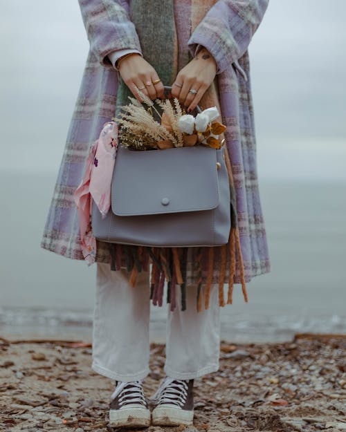 Free A woman in a coat holds a handbag with flowers, standing by the beach. Stock Photo