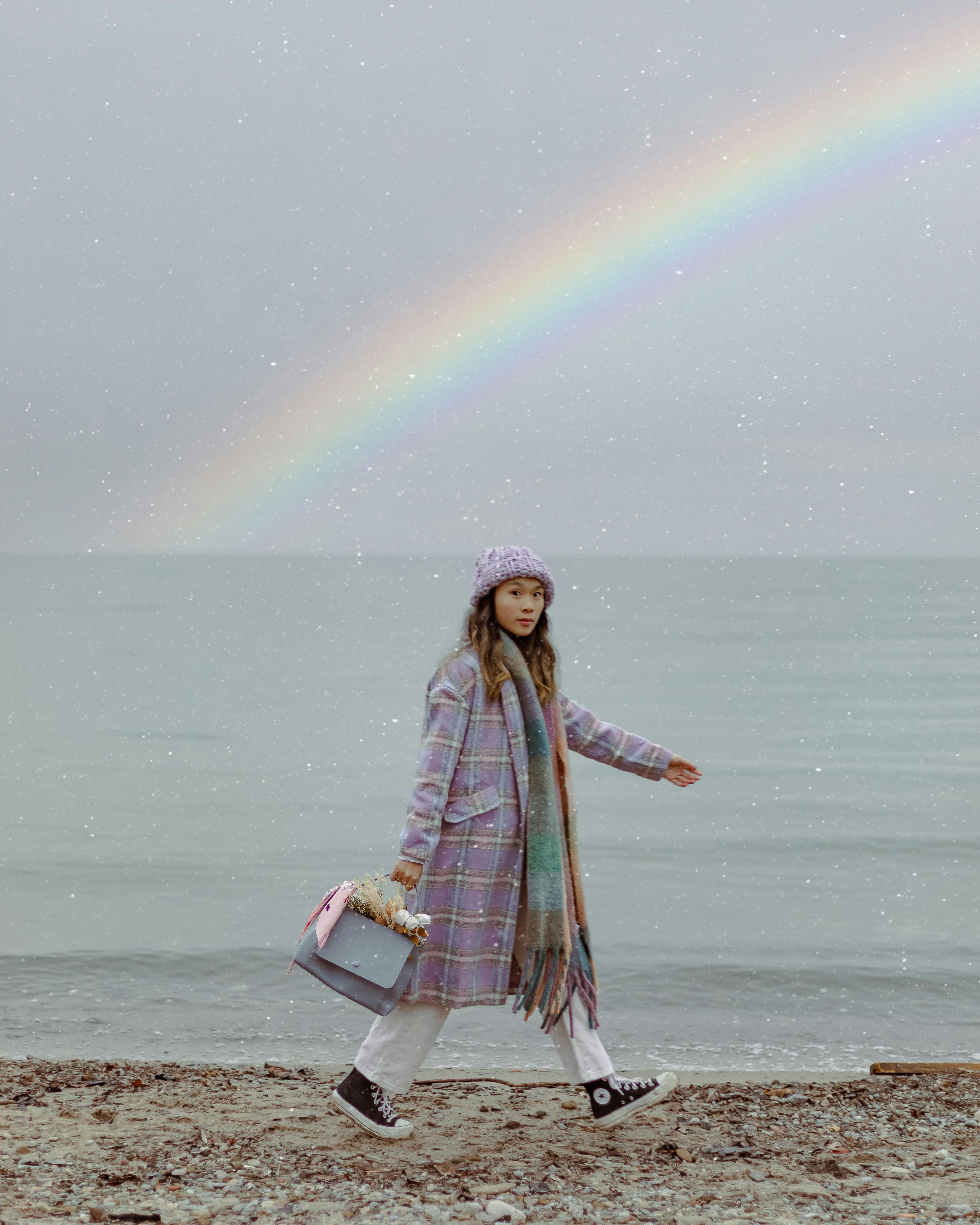 Young woman in plaid coat walking on the beach beneath a vibrant rainbow.