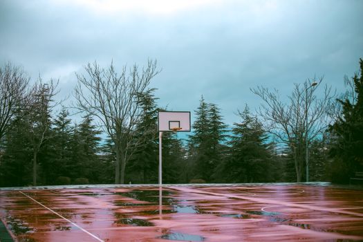 A scenic shot of a drenched basketball court after rain, surrounded by leafless trees.