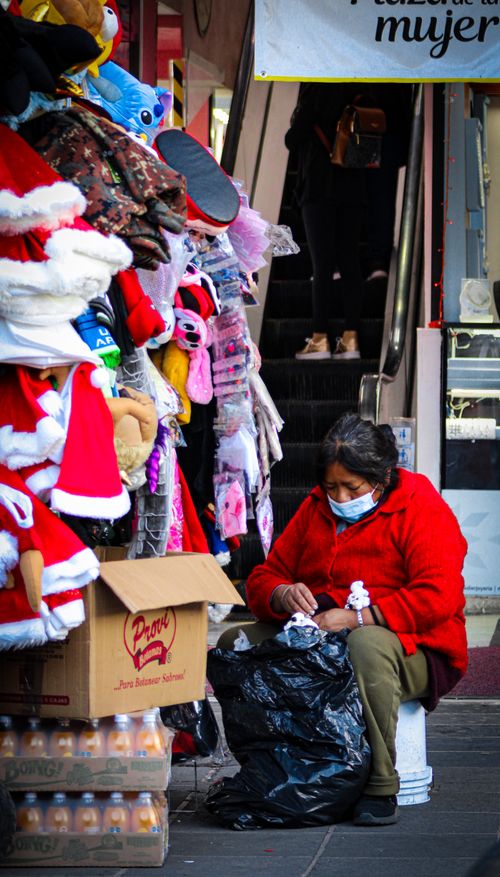 Free A street vendor in a red sweater wearing a face mask while working at a busy outdoor stall. Stock Photo