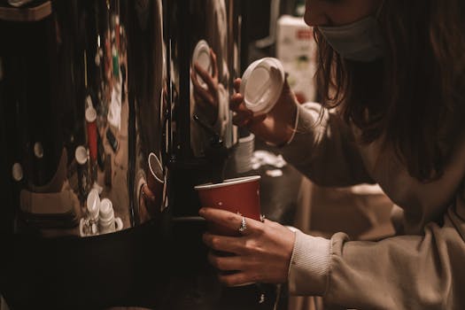 A woman pours coffee into a paper cup, capturing a cozy café atmosphere with warm lighting and reflections.