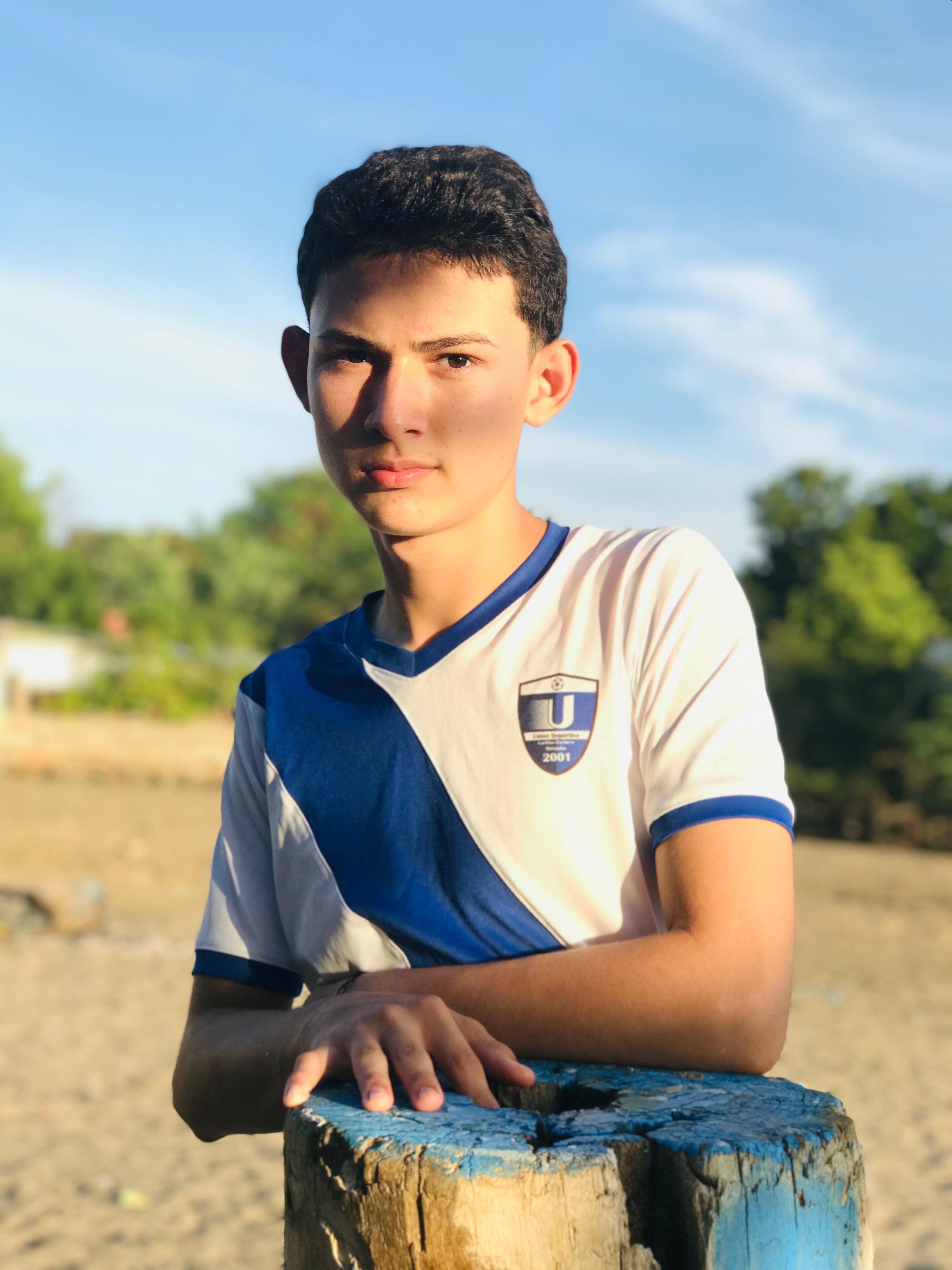 Portrait of a teenage boy outdoors wearing a white and blue t-shirt, leaning on a wooden post.