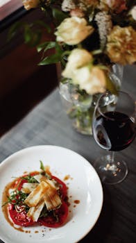 A gourmet meal elegantly styled with red wine and a floral centerpiece on a wooden table.