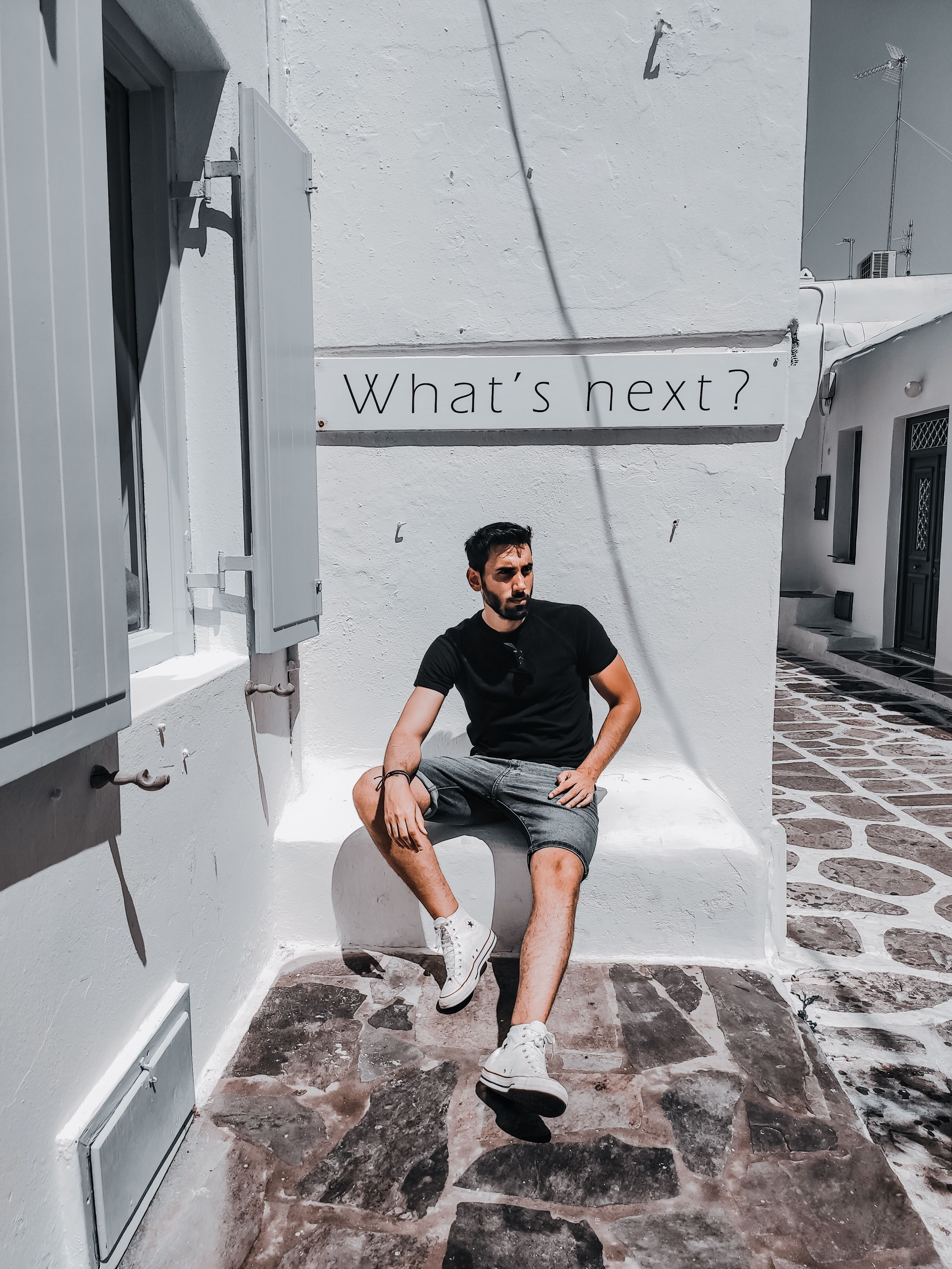 A man relaxes on a sunlit street in Mykonos, Greece, pondering the question 'What's next?'