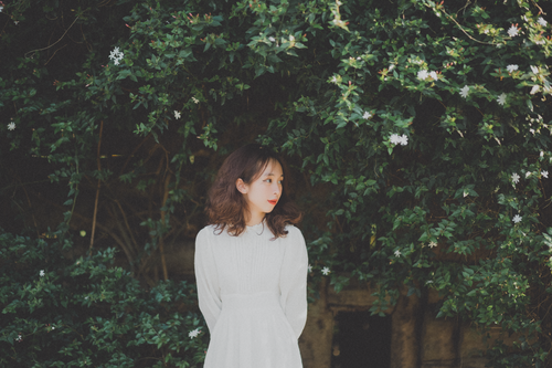 Free A woman in a white dress gazes sideways, surrounded by vibrant green leaves and delicate white flowers. Stock Photo