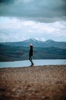 A moody, serene moment captured with a solitary figure walking along a lakeshore under a cloudy sky.