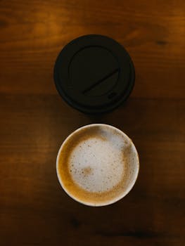 Aerial shot of a cappuccino with foam art next to a black cup lid on a wooden surface.
