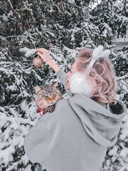 A woman decorates a snowy tree with a cat in a winter wonderland setting.
