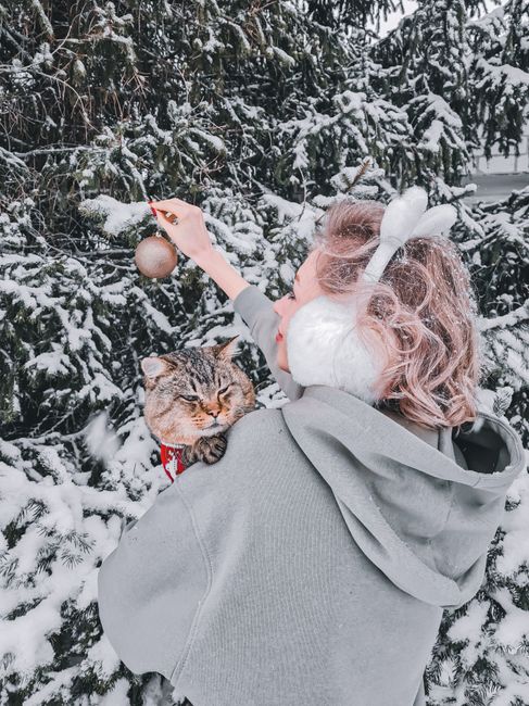 A woman decorates a snowy tree with a cat in a winter wonderland setting.