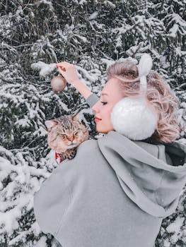 Woman with earmuffs holding a cat and decorating a snow-covered tree with a bauble.