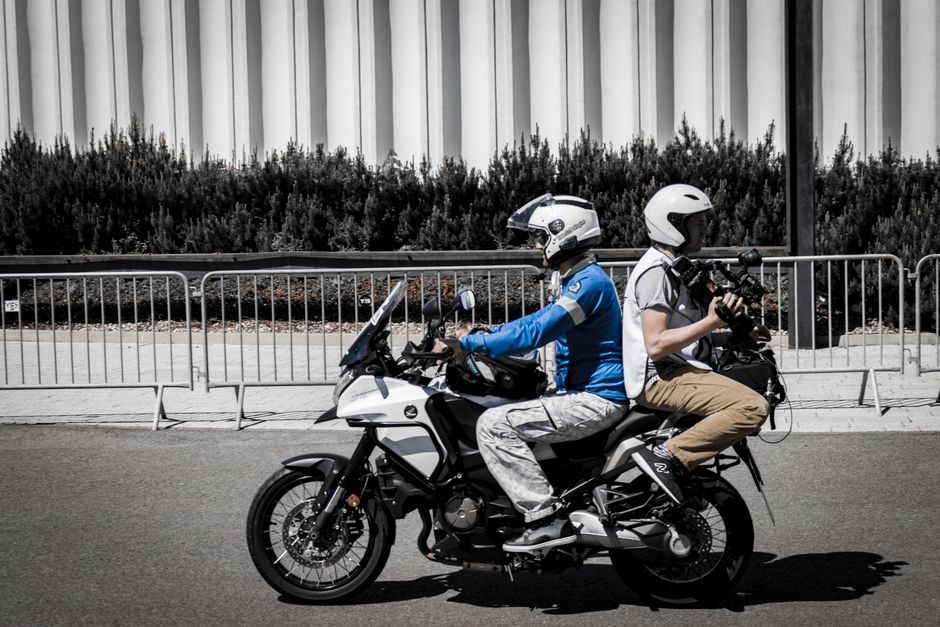 Two motorcyclists capturing video on a street with modern architecture.