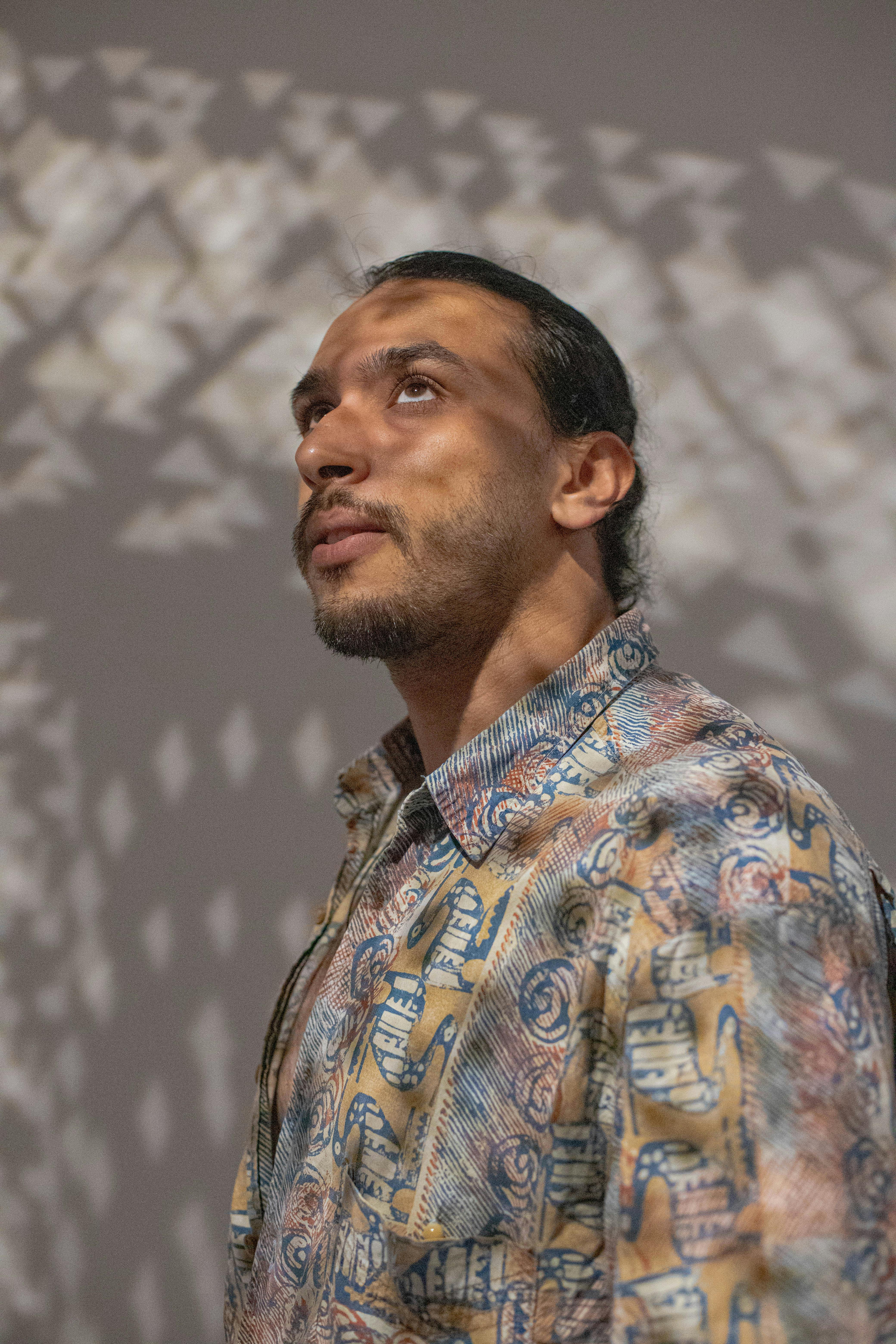 A man in a patterned shirt gazes upward amidst artistic shadow play on a wall.