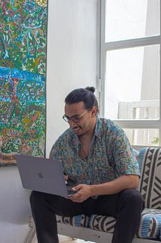 Young man smiling while using a laptop in a stylish indoor setting with artistic decor.