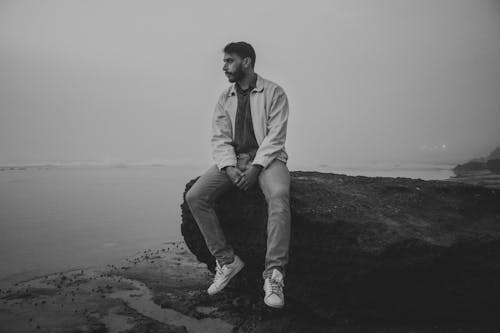 Grayscale Photo of Man Sitting on Rock Formation near Ocean