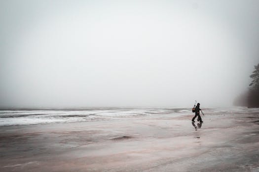 Lonely fisherman with equipment walking on a foggy, reflective seashore, emphasizing solitude.
