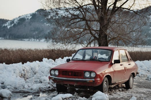 Vintage red car parked on a snowy roadside with winter mountains in the background.