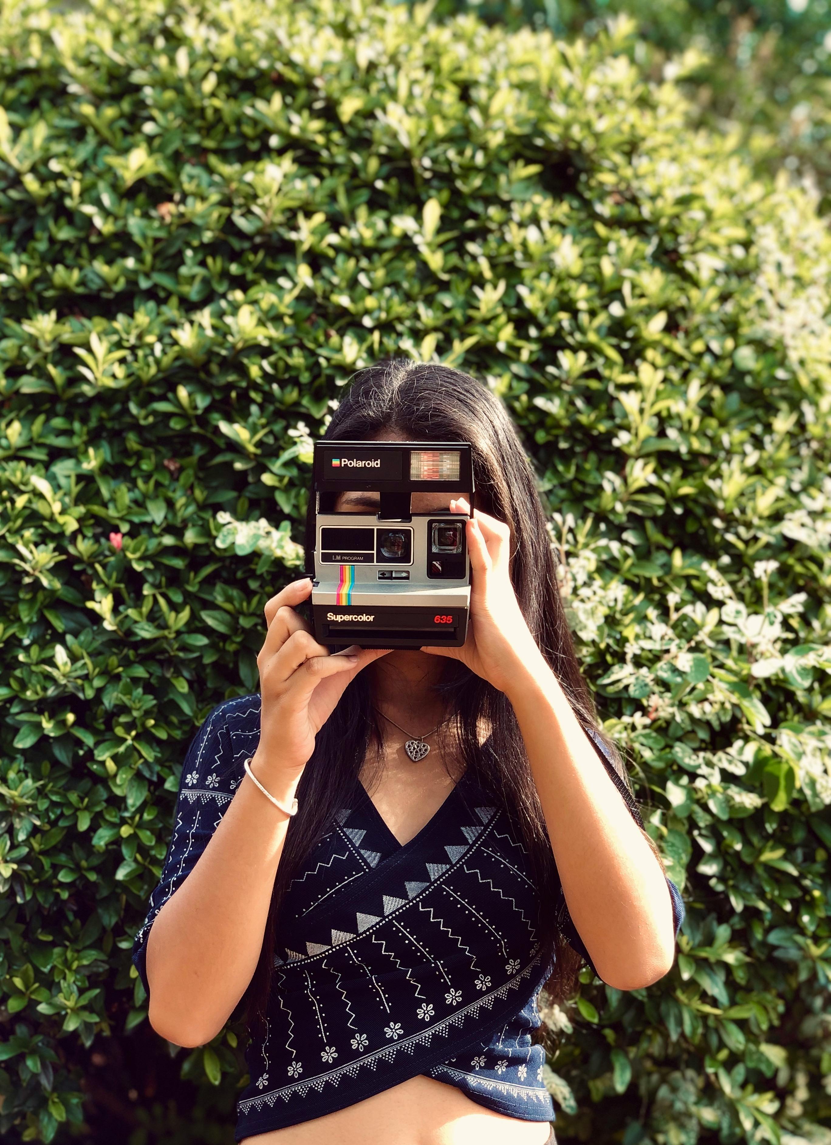 Woman holding a vintage Polaroid camera against a lush green backdrop, capturing a moment in nature.