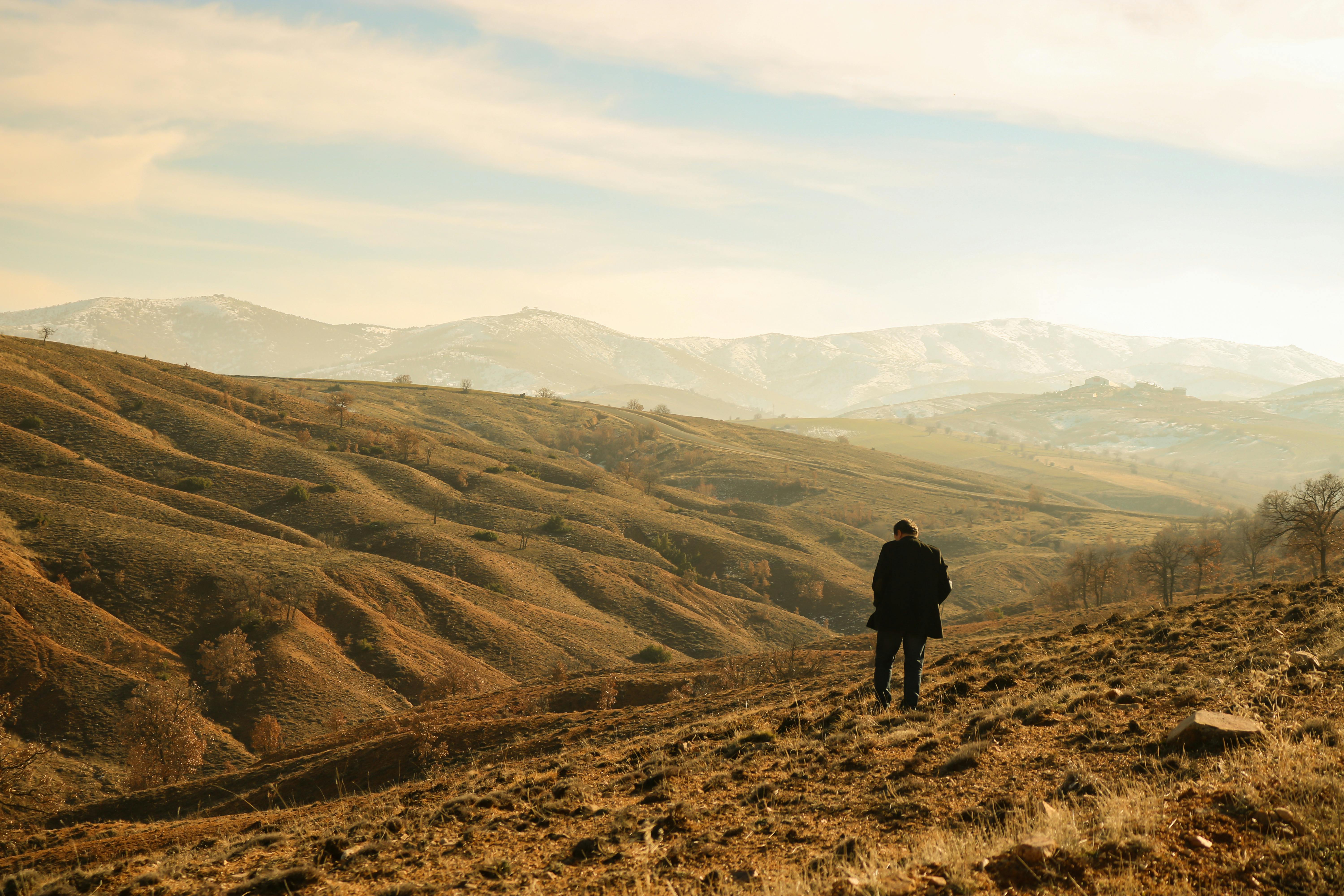 A lone person standing amidst rolling hills and distant mountains under a clear sky.