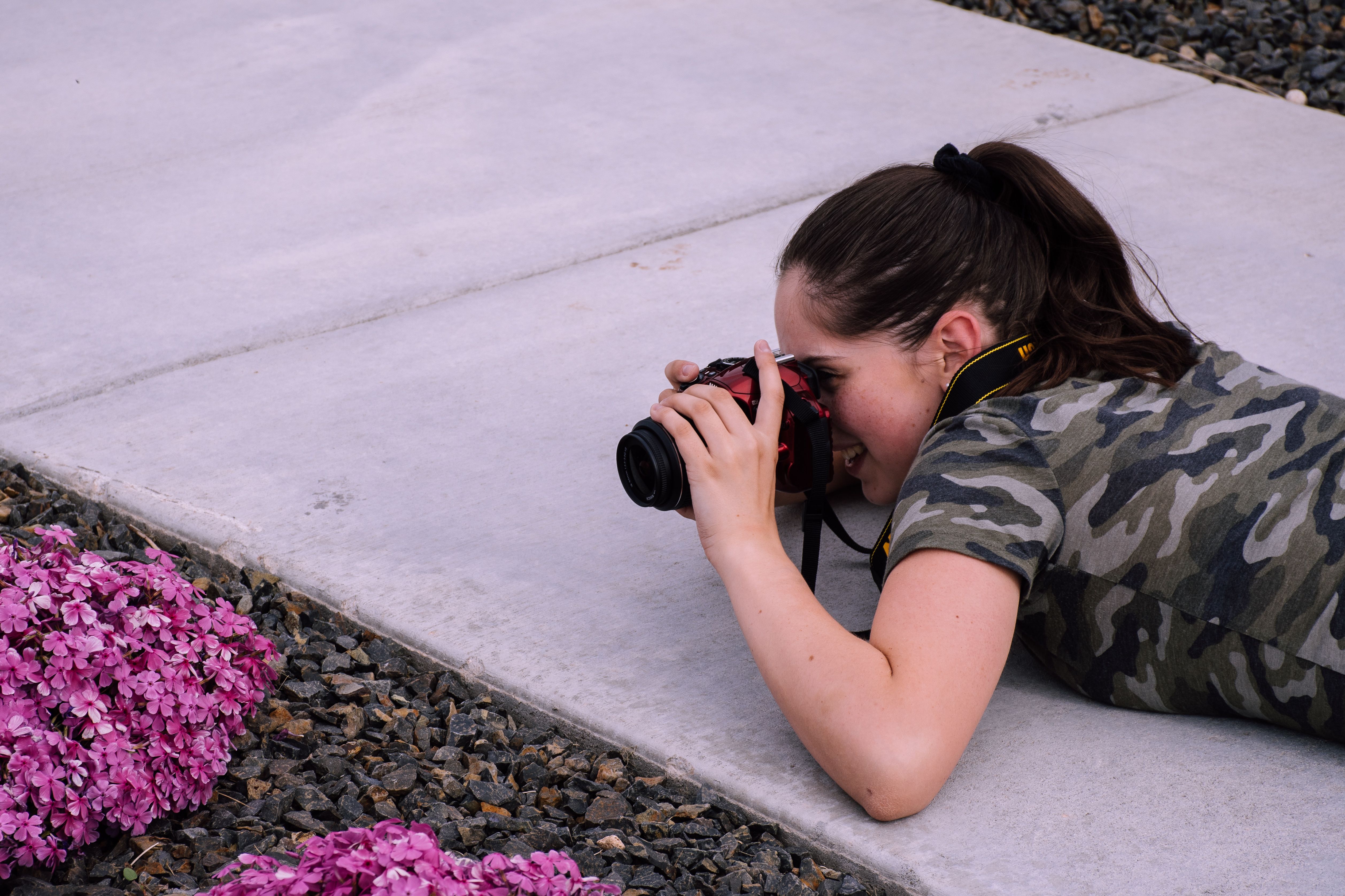 Young woman photographer in camouflage shirt capturing flowers on pavement.