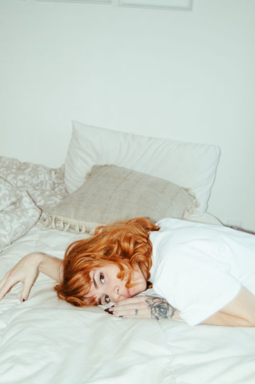Free A woman with tattoos and red hair relaxes on a bed, wearing a crew neck t-shirt, in a modern bedroom setting. Stock Photo