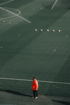 A lone man in a red jacket standing on a football field with soccer balls and sprinklers.