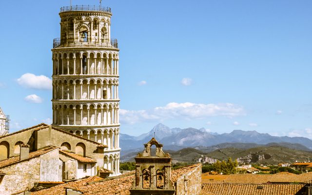 Photo: Russell Butcher on Pexels Leaning Tower of Pisa in Italy framed by rooftops and distant mountains under a clear blue sky.