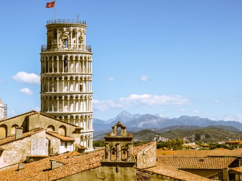 Free Leaning Tower of Pisa in Italy framed by rooftops and distant mountains under a clear blue sky. Stock Photo