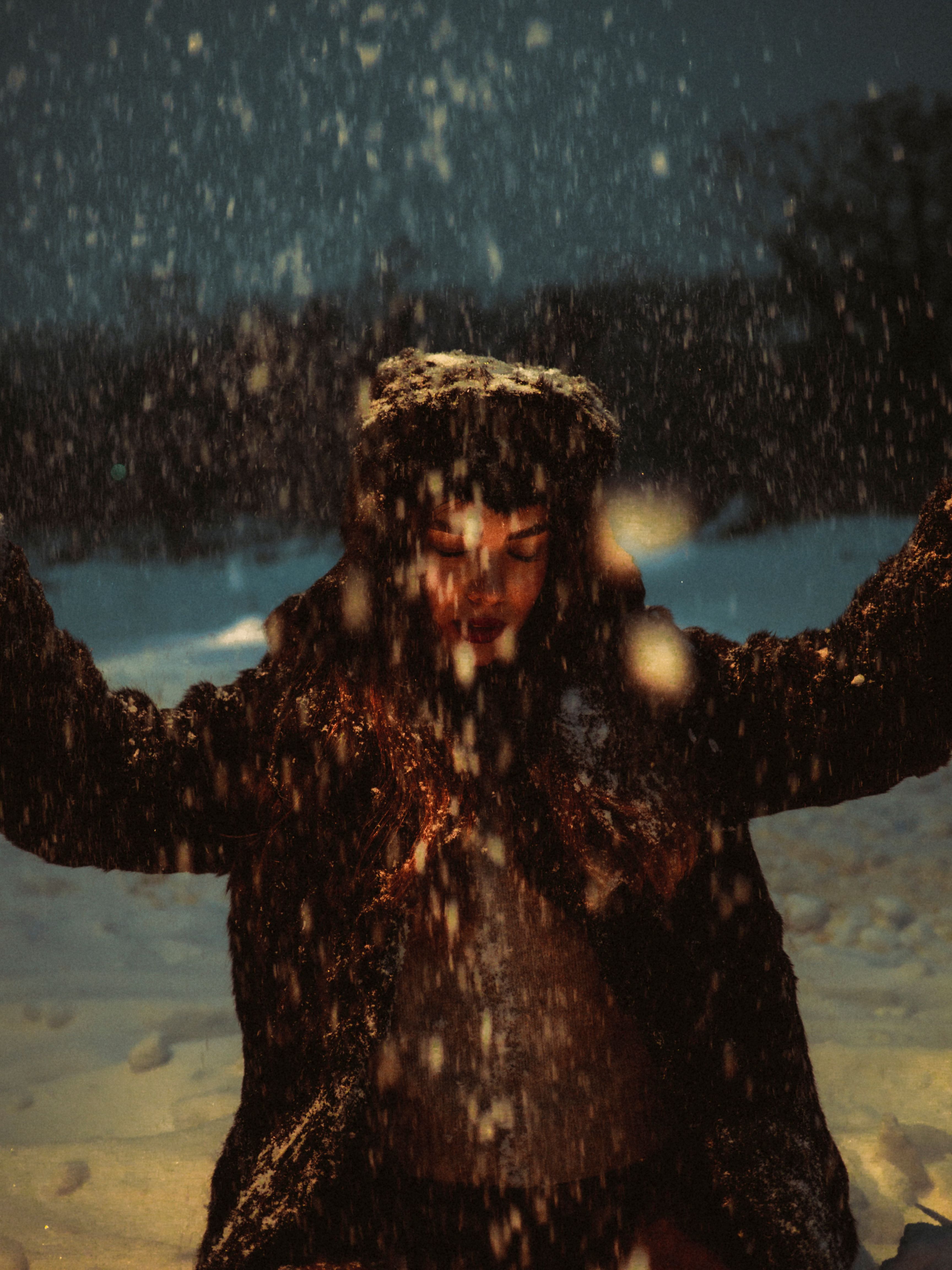 A woman dressed warmly experiences the joy of snowfall during a winter evening.