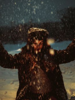 A woman dressed warmly experiences the joy of snowfall during a winter evening.