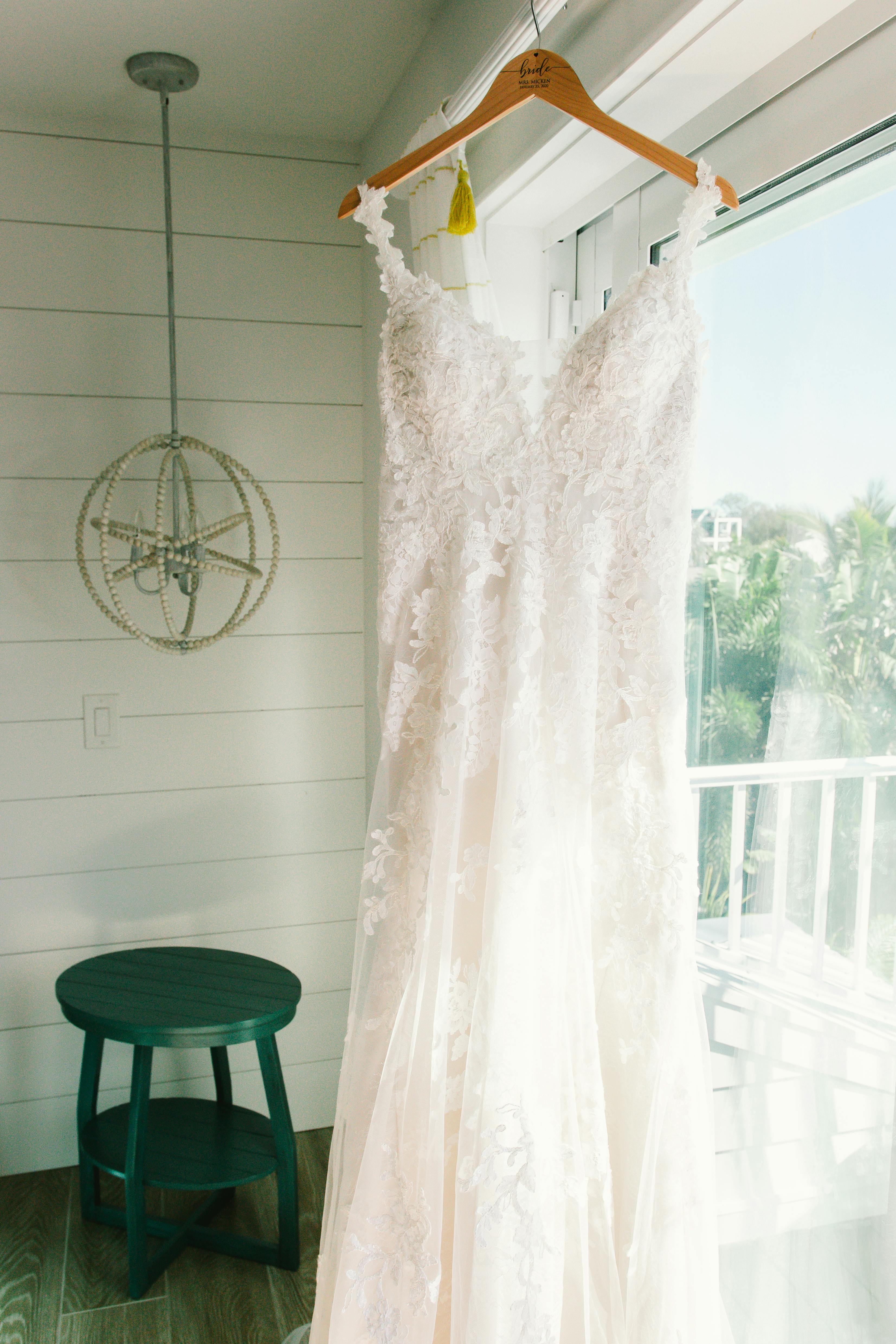 White lace wedding gown hanging near a bright window in a sunlit room, capturing the elegance of bridal fashion.