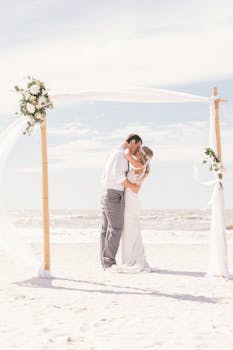 Newlyweds share a romantic kiss under a floral arch on a sandy beach in Florida.