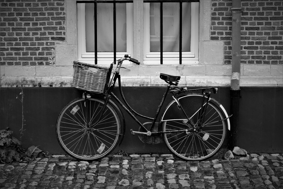 Monochrome image of a vintage bicycle with a basket against a cobblestone street and brick facade.