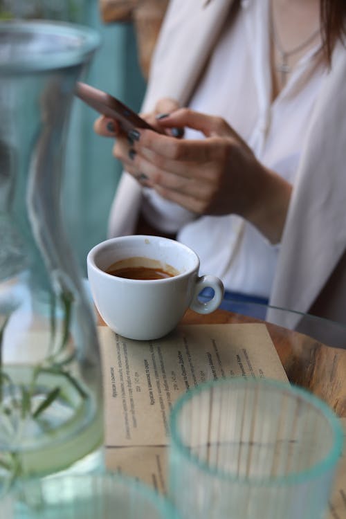 Person Holding a Smart Phone Over a White Ceramic Mug With Coffee