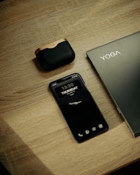 Flat lay of smartphone, laptop, and earbuds on a wooden desk, perfect for business and tech themes.