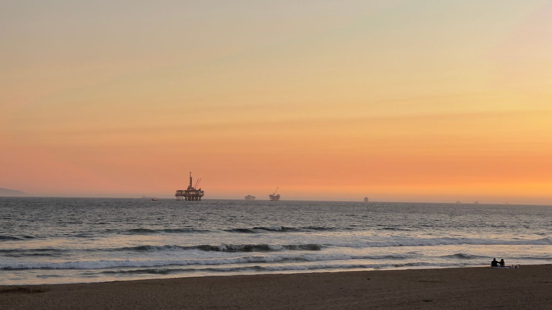 Serene beach view with distant oil rigs under a vibrant sunset sky.