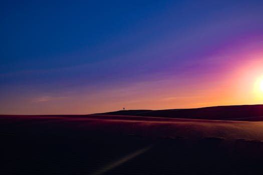 Vibrant sunset over rolling desert dunes with silhouetted figures on the horizon.