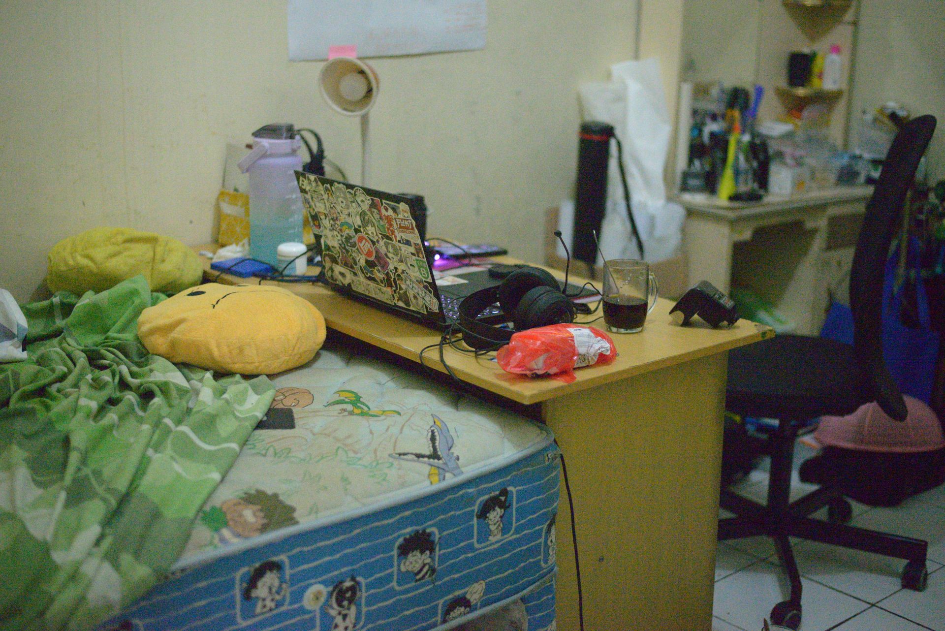 A cluttered workspace with a laptop, headset, and bed in a cozy room setting.